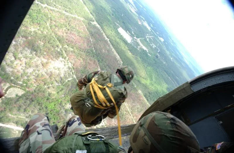 View from inside a military aircraft, paratrooper preparing to exit the door over forested terrain.